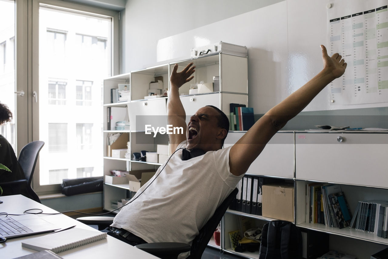 Happy male entrepreneur screaming with arms raised while sitting at office