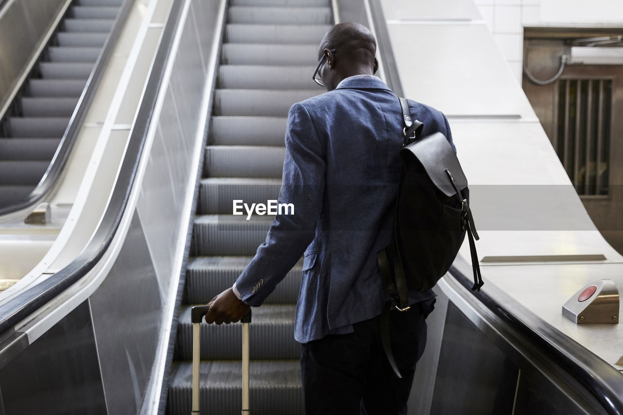 Rear view of businessman standing with luggage moving up on escalator at subway station