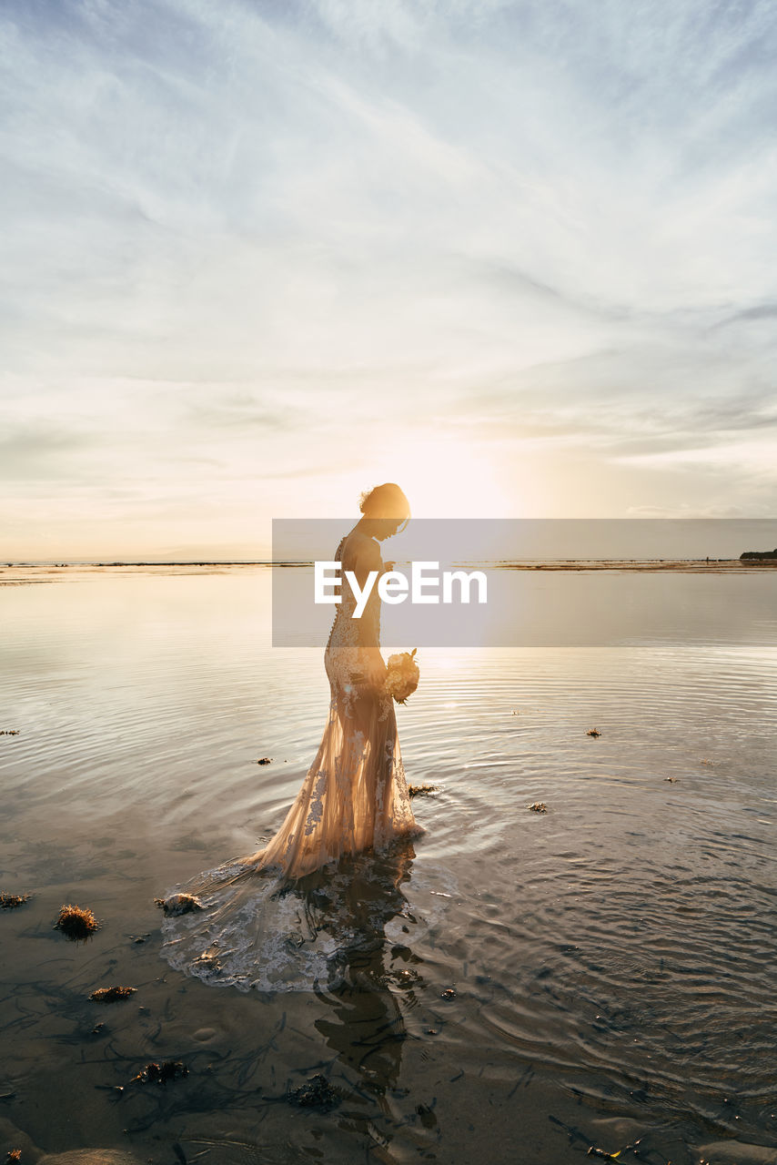 Side view of bride standing at beach against sky during sunset