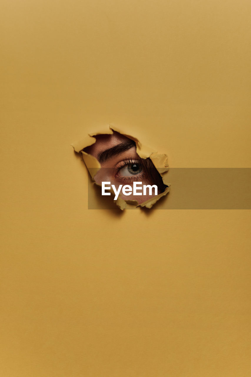 Eye of unrecognizable female model looking through ripped hole in yellow paper person
