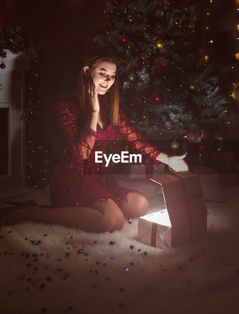 Young woman sitting on carpet by illuminated gift box and christmas tree at home