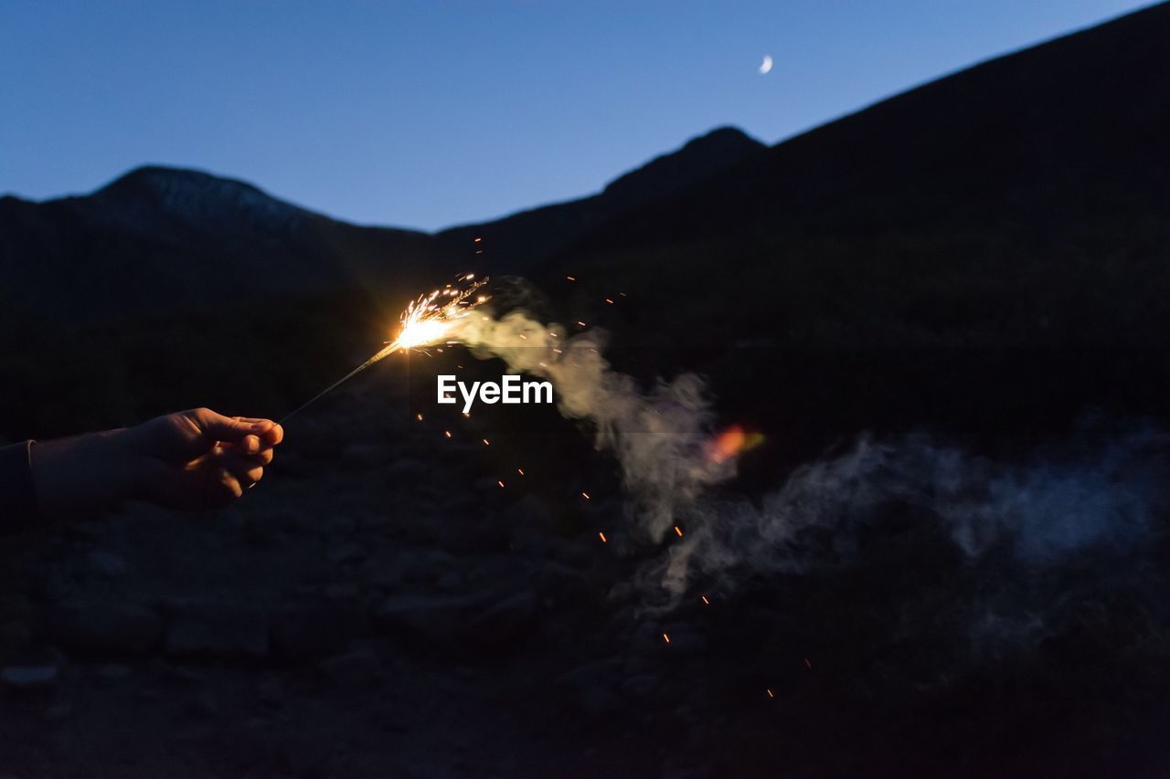 Cropped hand of person holding sparkler against mountain at night