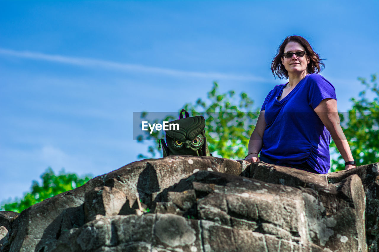 Low angle view of mature woman sitting on rock against blue sky