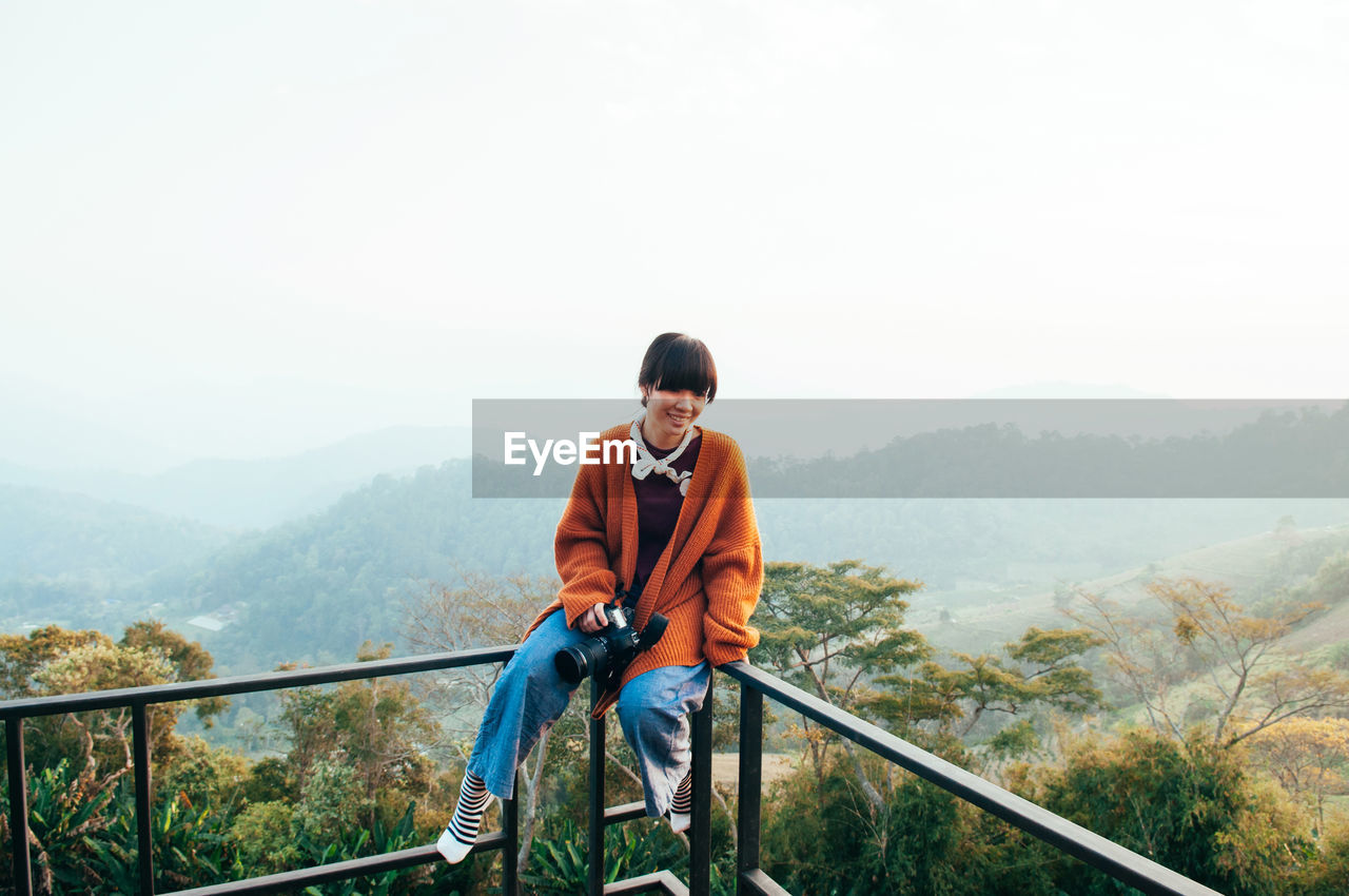 Young asian woman wearing casual clothes holding camera sit on fence at mountain top