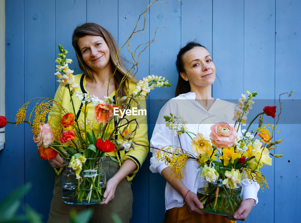 Two young women with beautiful bouquets of peonies in glass vases against the blue background