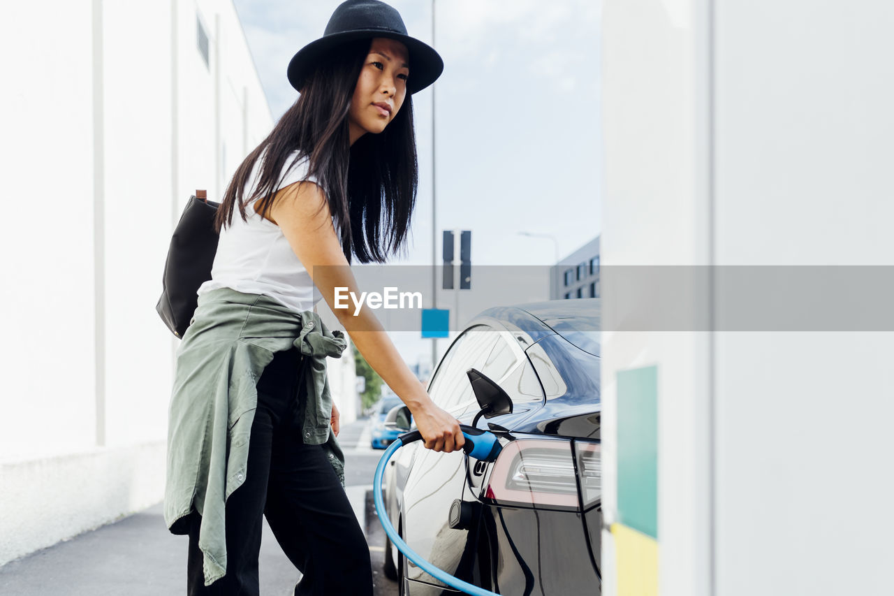 Young woman wearing hat charging electric car at vehicle station