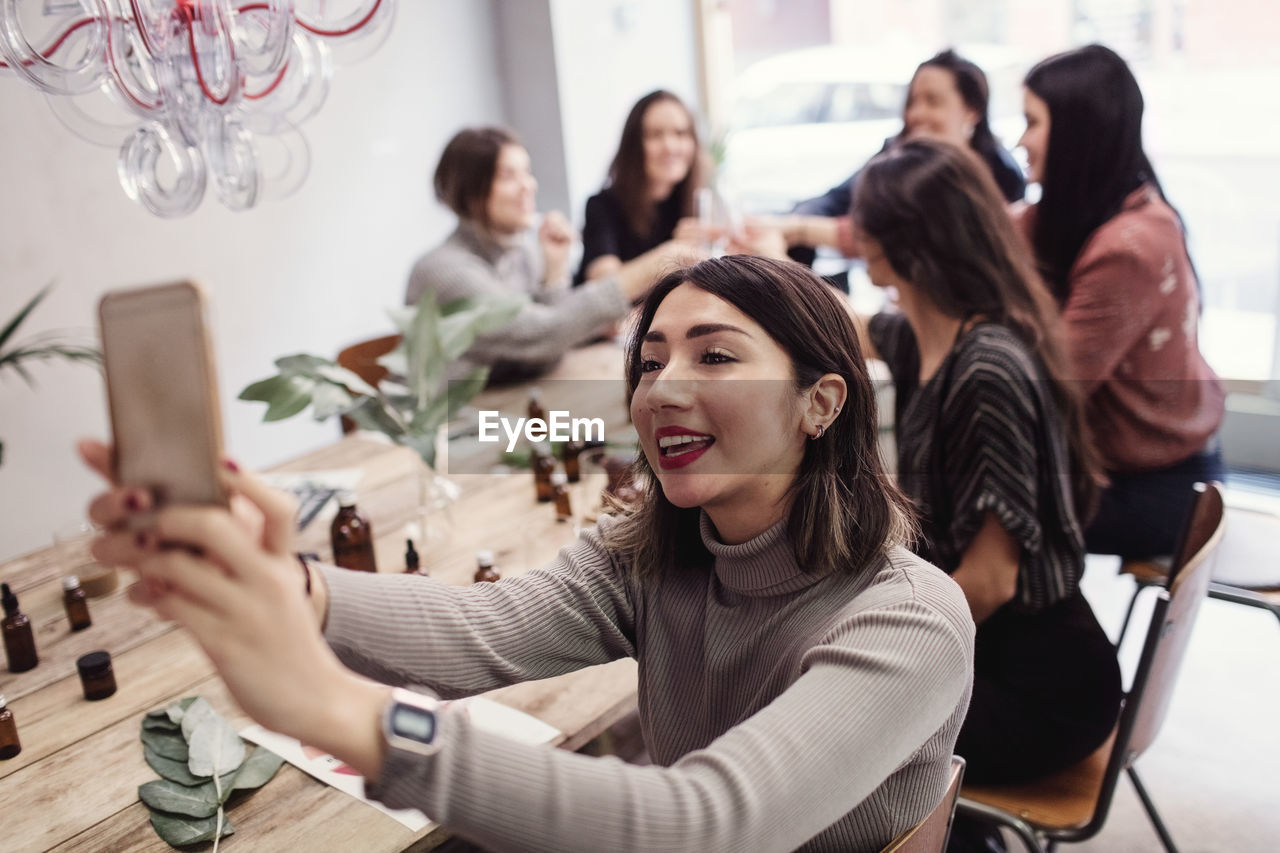 Smiling young woman taking selfie with female colleagues sitting at table in perfume workshop