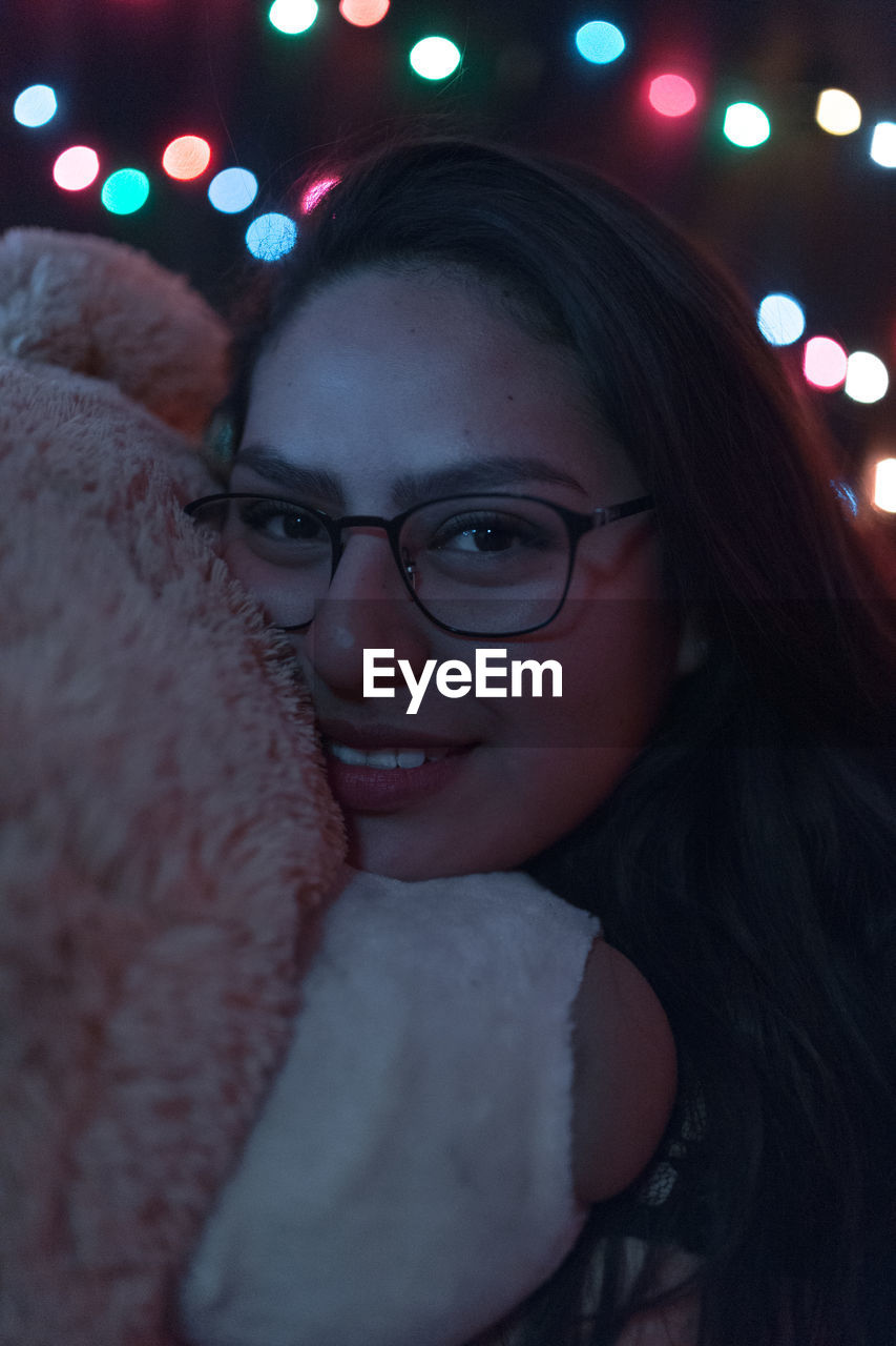 Close-up portrait of young woman with teddy bear against illuminated lights