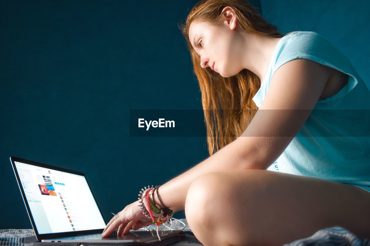 Woman in a bed working from home using laptop against blue wall