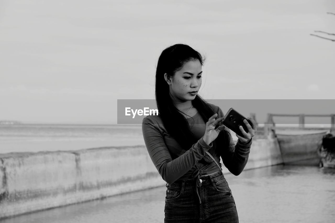 Young woman using mobile phone while standing against sky