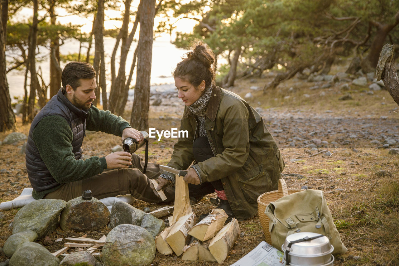 Man pouring drink while woman with firewood at campsite in forest