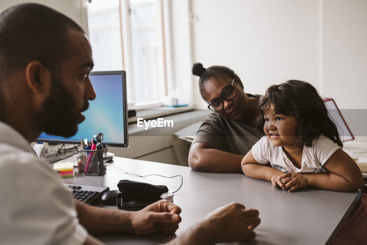 Male pediatrician talking to girl sitting with mother at desk