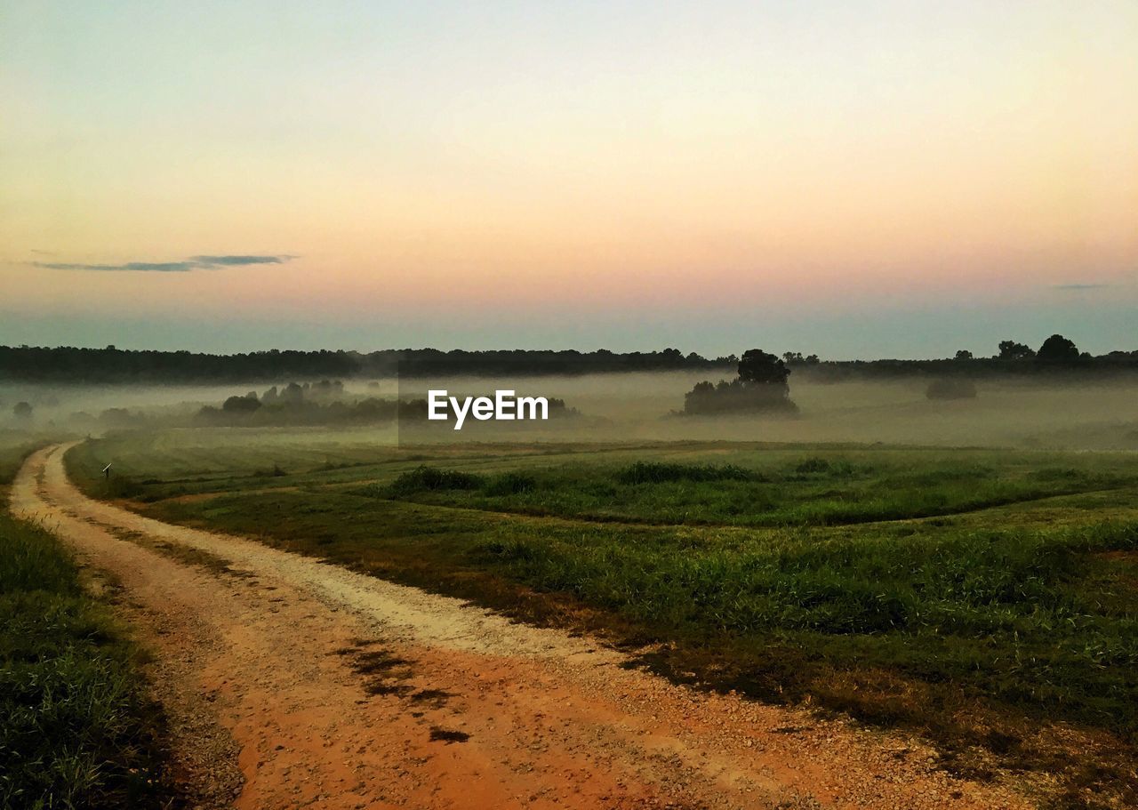 Dirt road amidst field against sky during sunset