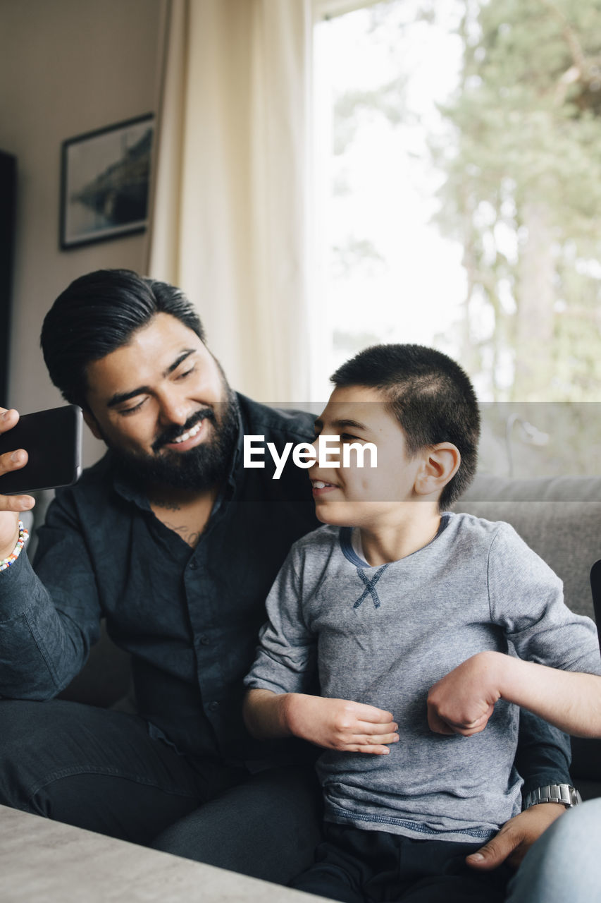 Smiling father holding mobile phone looking at autistic son while sitting on sofa in living room
