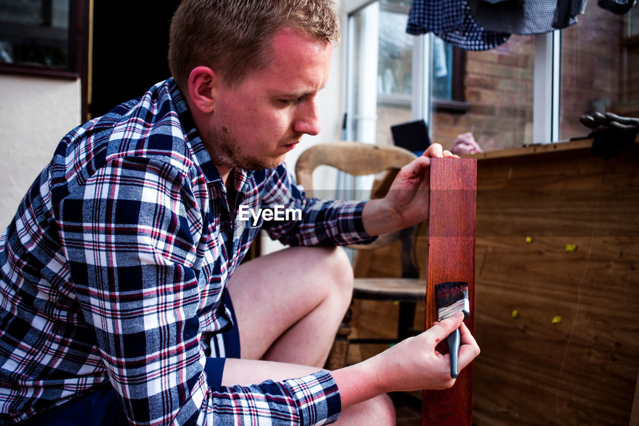 Side view of young man painting wooden plank while crouching at home
