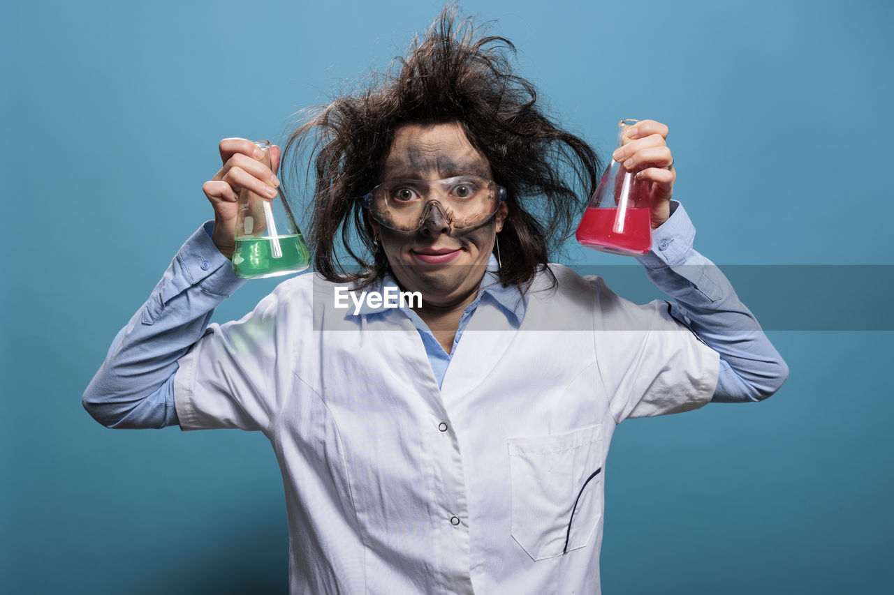 studio shot, one person, colored background, adult, indoors, science, person, blue, clothing, portrait, scientist, human face, blue background, occupation, education, holding, waist up, research, chemistry, looking at camera, front view, healthcare and medicine, nose, men, scientific experiment, human mouth, lab coat, human head, humor, emotion, laboratory, fun, eyeglasses, glasses, professional occupation, hand, bottle, arm