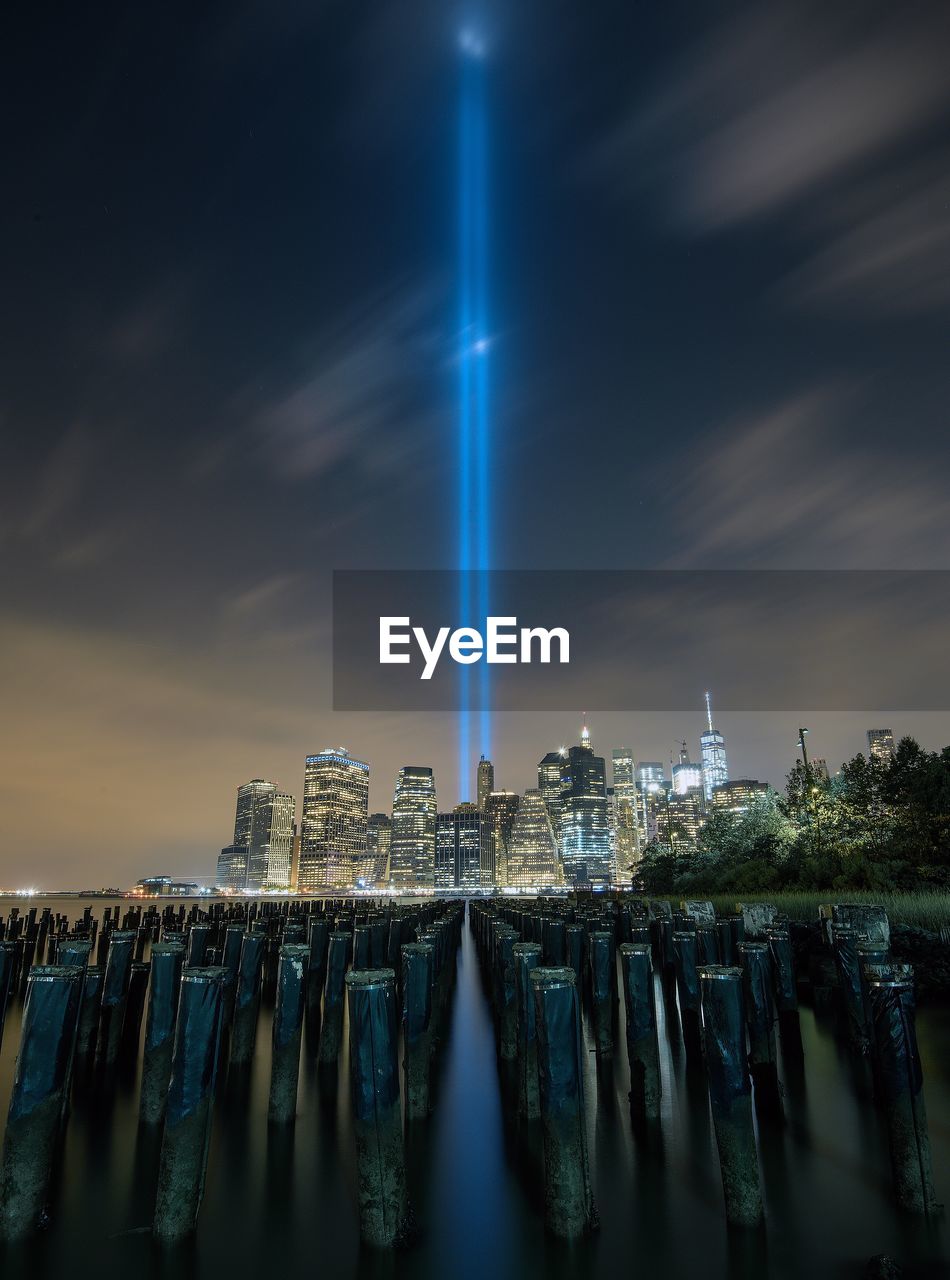 Wooden post in east river against light beams symbolizing world trade center in city against sky at night