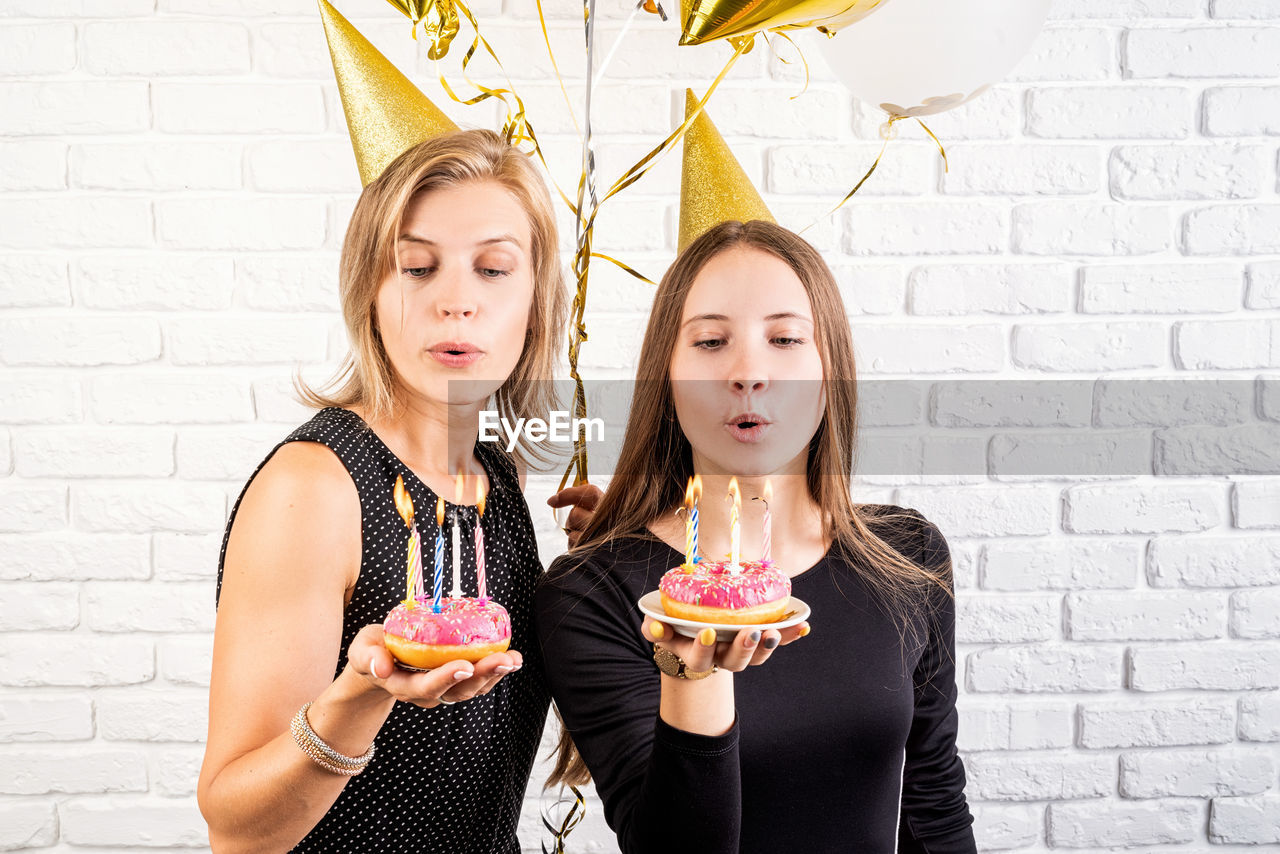 Two smiling young women or sisters in birthday hats celebrating birthday holding donuts with candles
