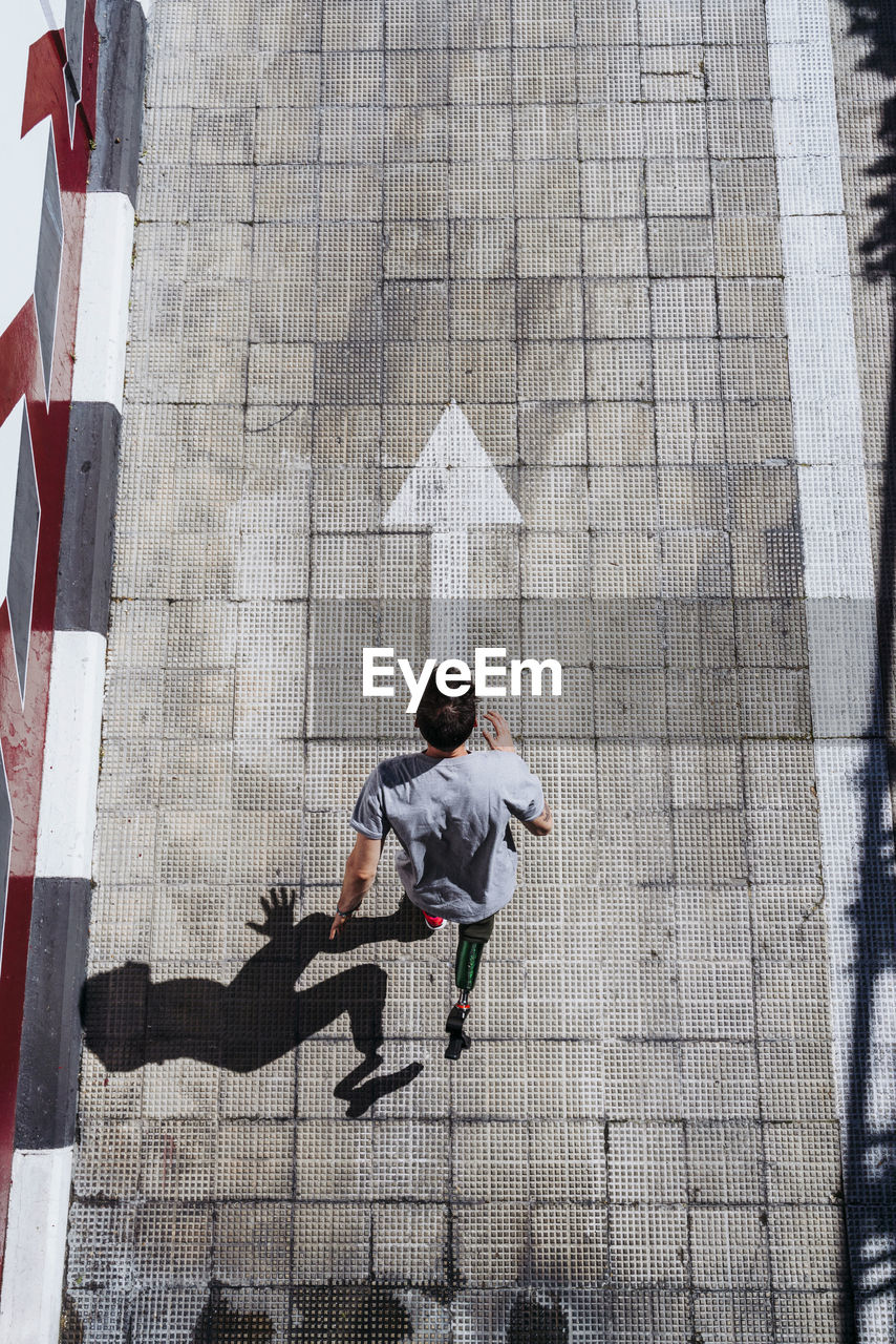 From above young guy with leg prosthesis running up paved slope against shabby building during workout on city street