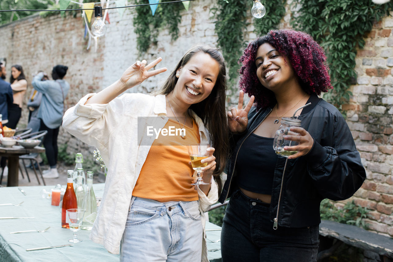 Portrait of happy female friends with drinks showing peace sign during dinner party in back yard