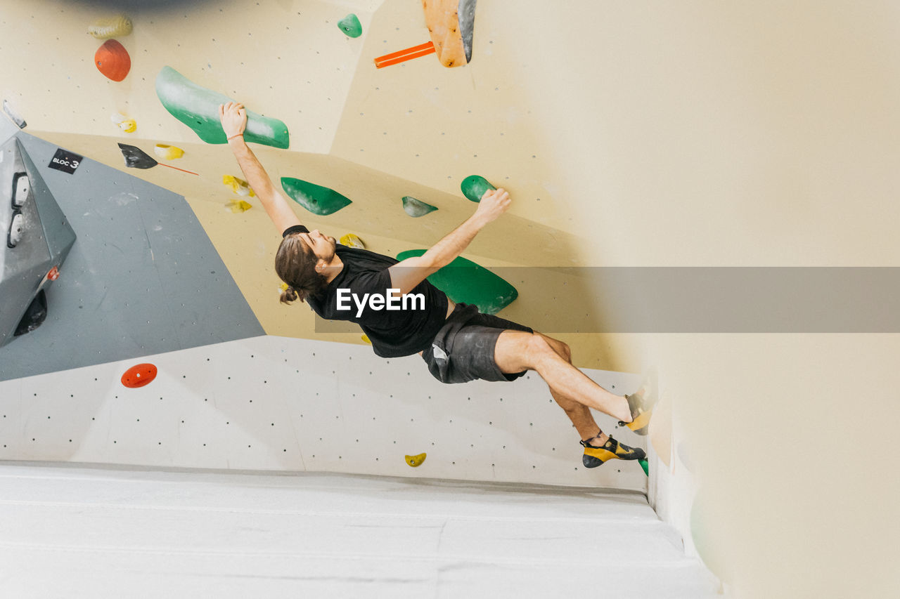 Full body side view of sportive male alpinist climbing wall with colorful grips during intense training in light modern gym