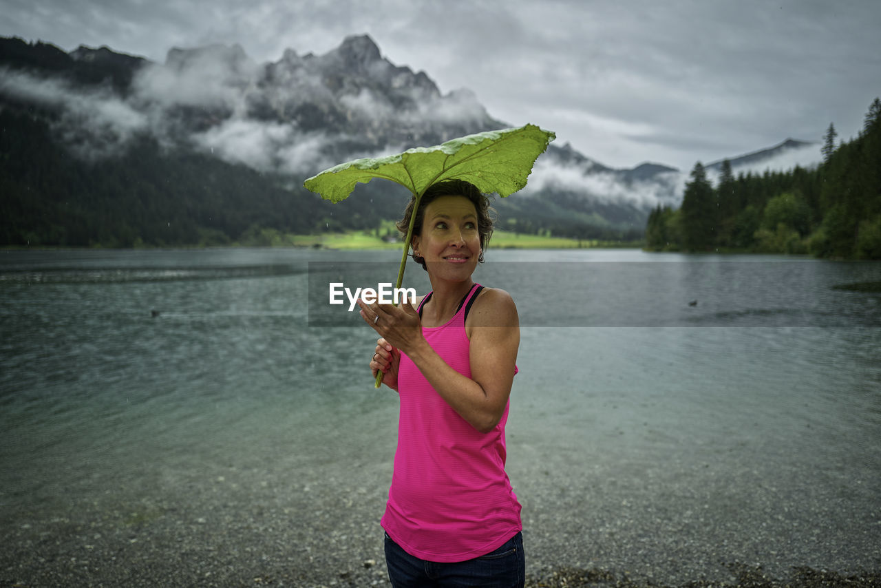 Smiling woman holding green leaf while standing at haldensee lakeshore during rainy season