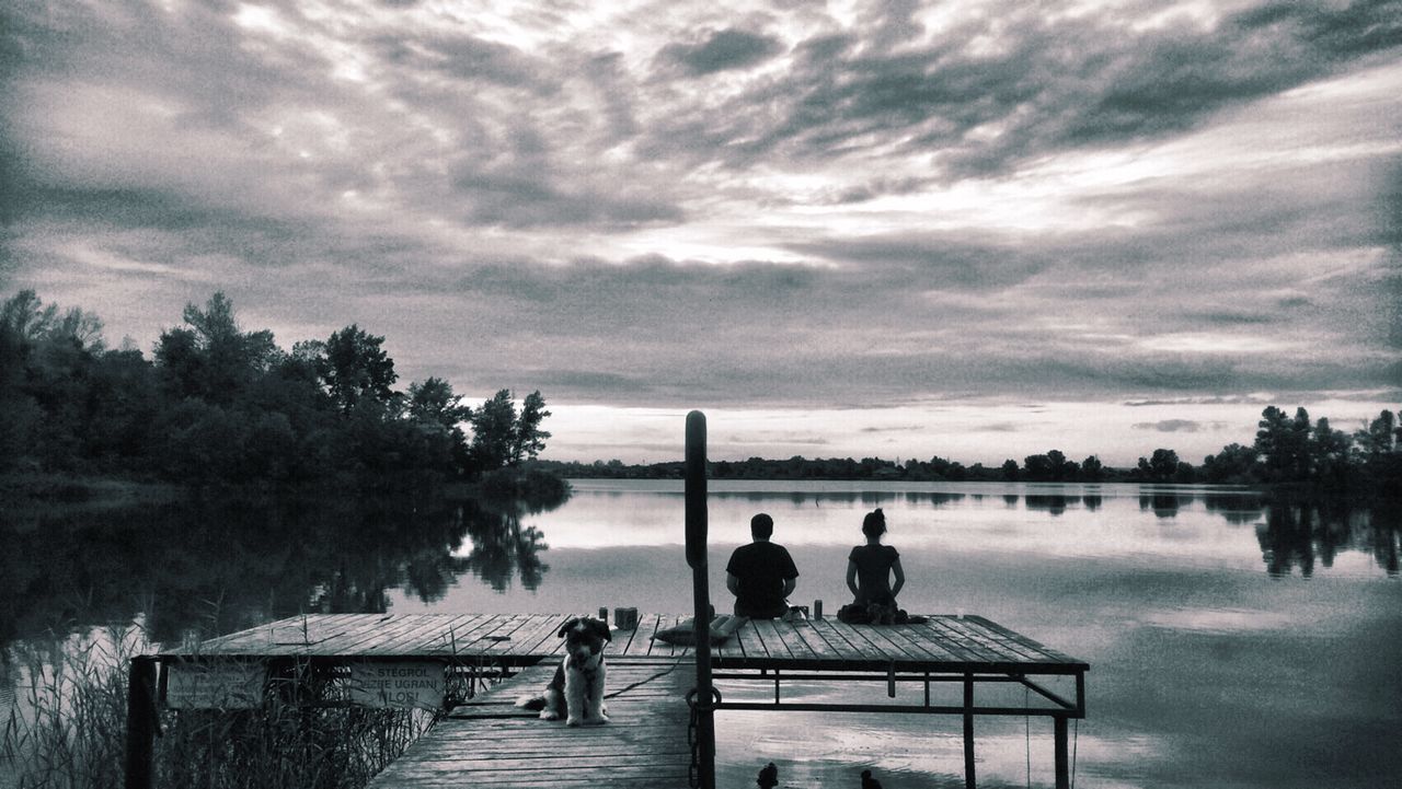 Rear view of man and woman with dog in foreground sitting on pier over lake