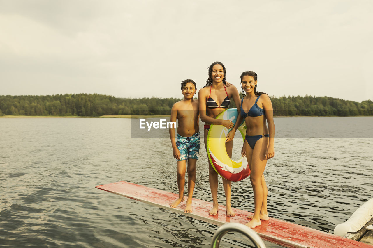 Smiling girl with inflatable ring standing with siblings on diving board at lake