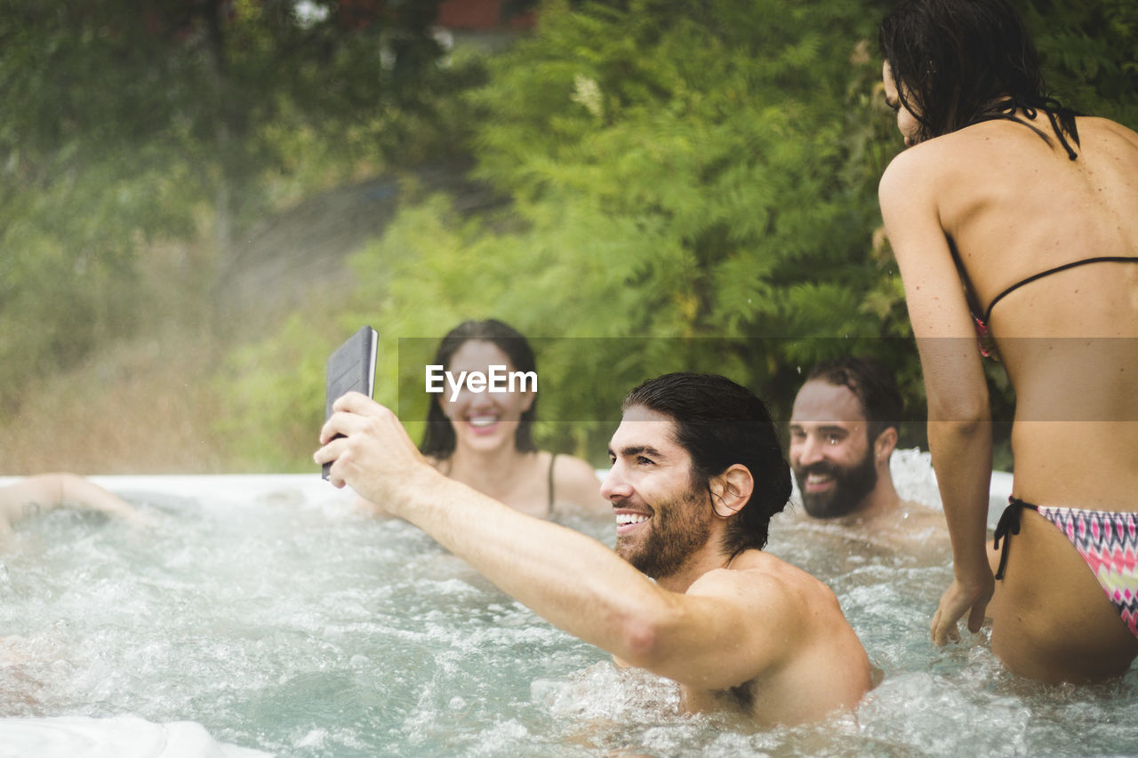 Young man taking selfie through mobile phone while friends enjoying in hot tub during weekend getaway