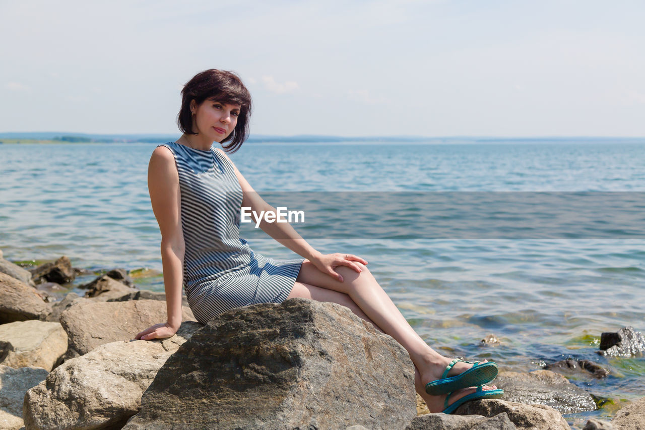 Young woman sitting on rock by sea against sky
