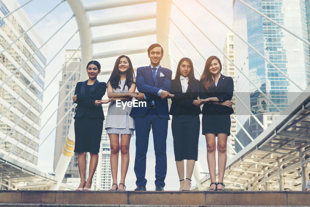 Portrait of smiling young colleagues with arms crossed holding hands on footpath against buildings in city