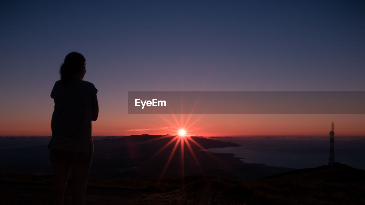 Rear view of silhouette woman standing at beach against clear sky during sunset