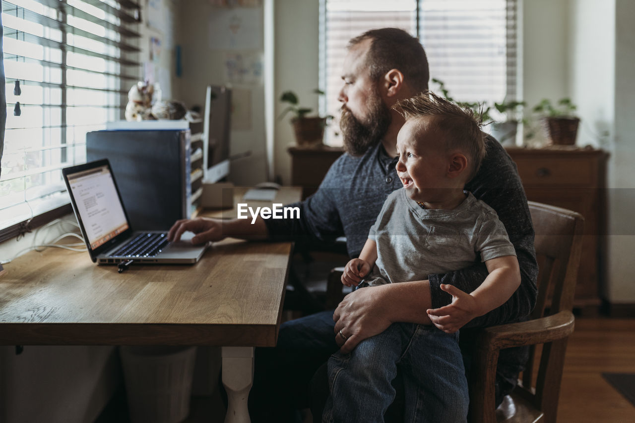 Dad working from home with one year old boy crying in his lap