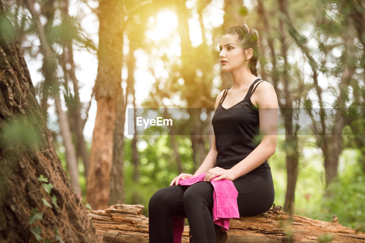 young woman looking away while standing against tree trunk in forest