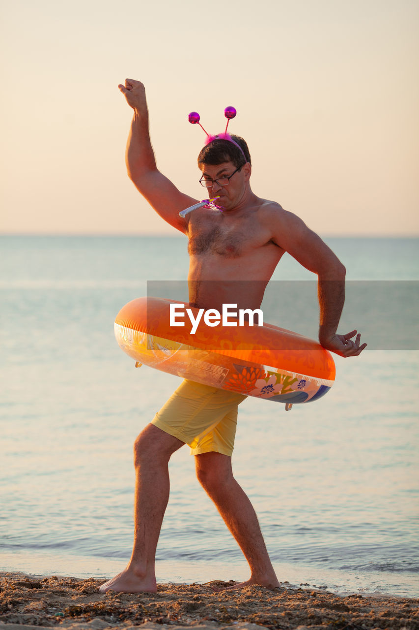 Full length of shirtless man with inflatable ring standing at beach during sunset