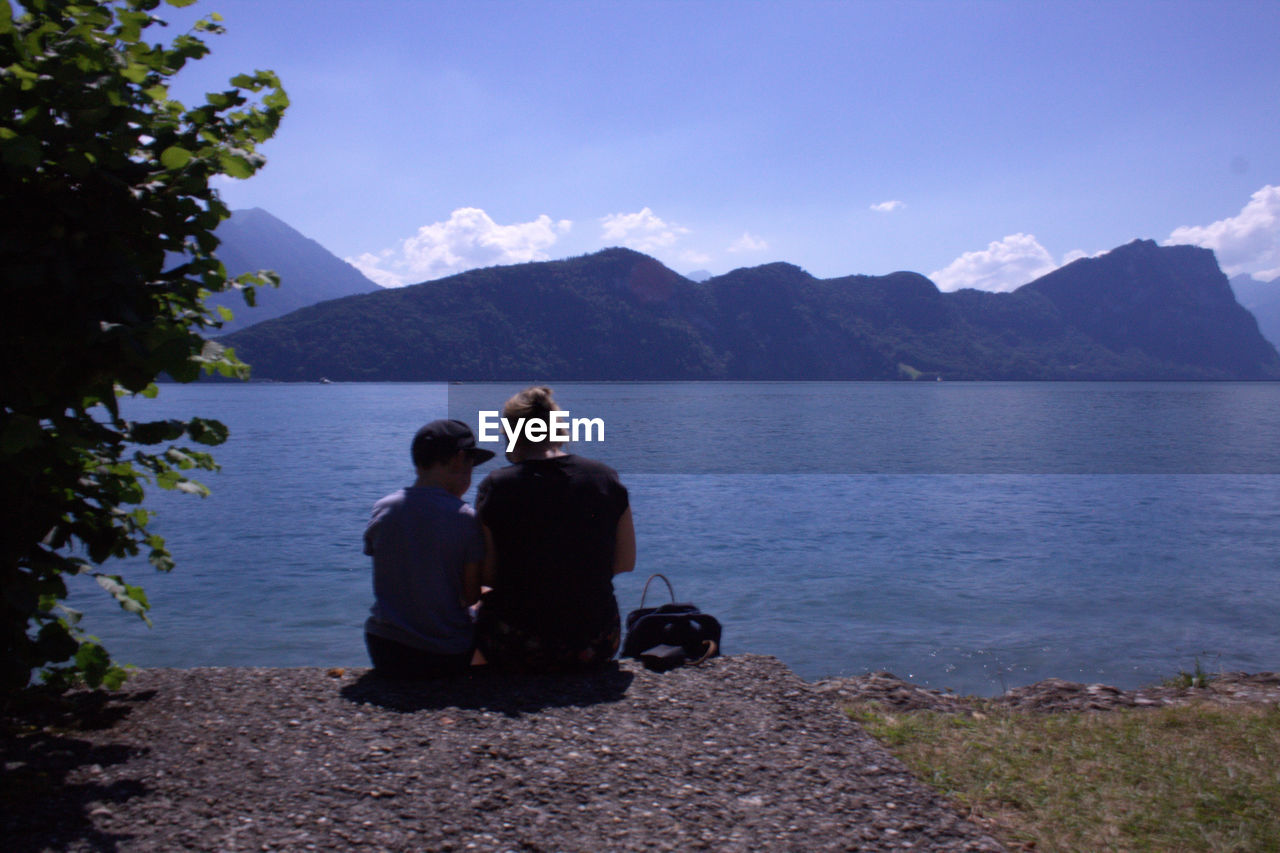 PEOPLE SITTING BY LAKE AGAINST MOUNTAINS