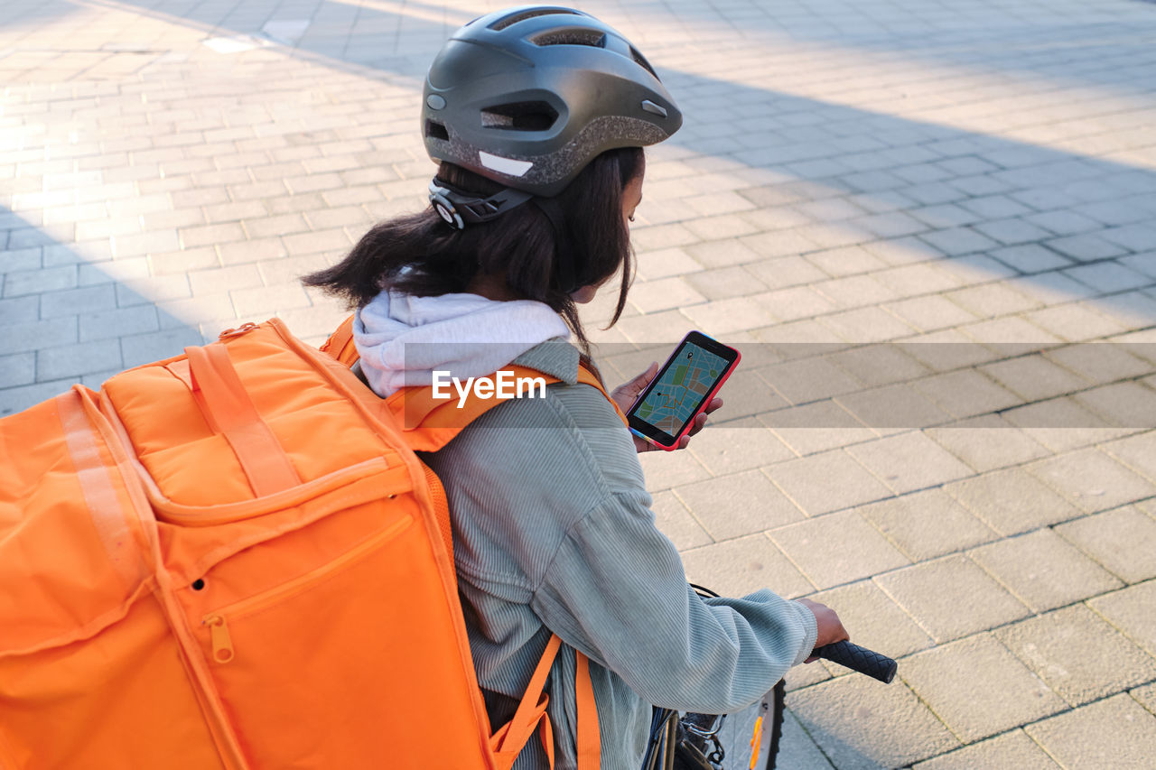 African american female cyclist with thermo backpack standing on paved sidewalk with bicycle while checking location information on cellphone
