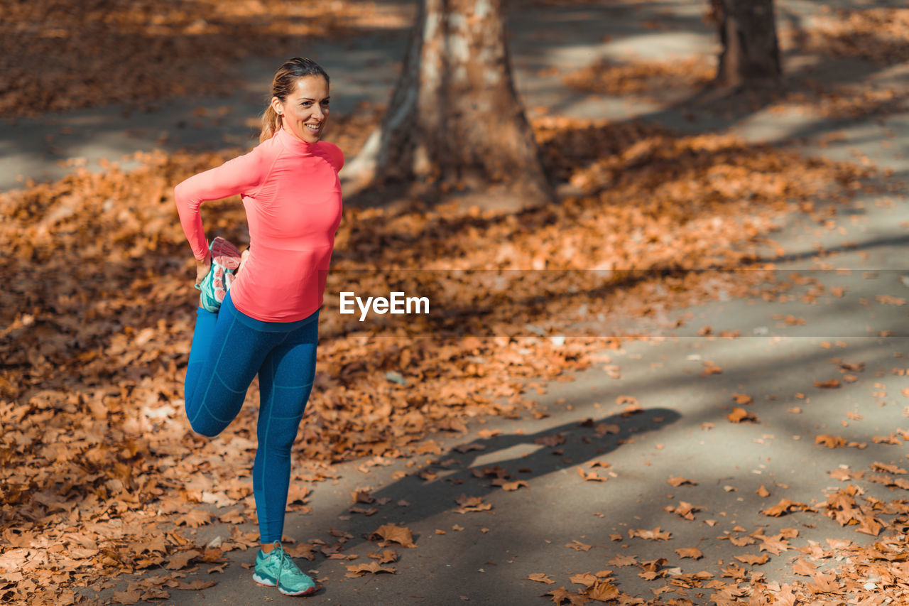 Smiling woman stretching while standing on footpath in park during autumn