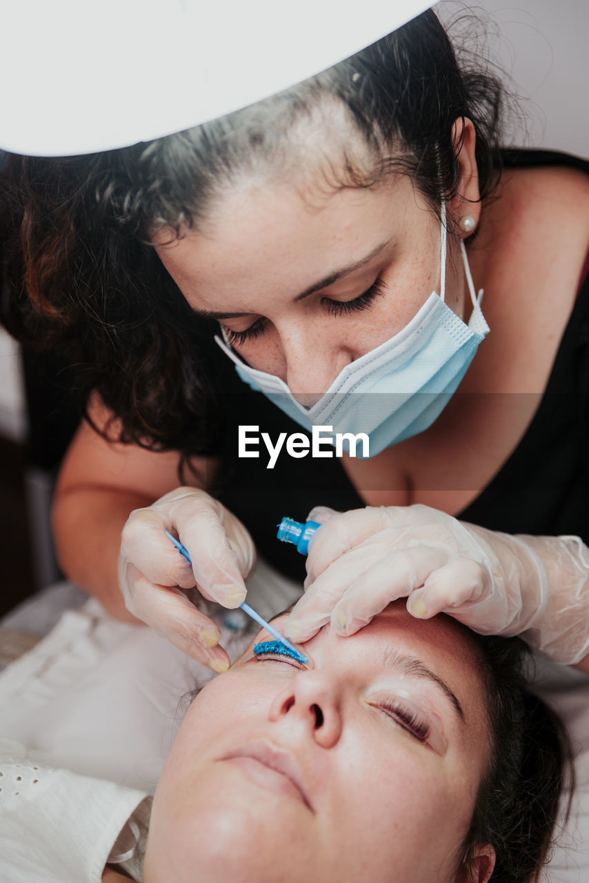 From above cosmetician in latex gloves applying blue chemical solution on eyelid of female client during eyelashes lifting procedure in salon