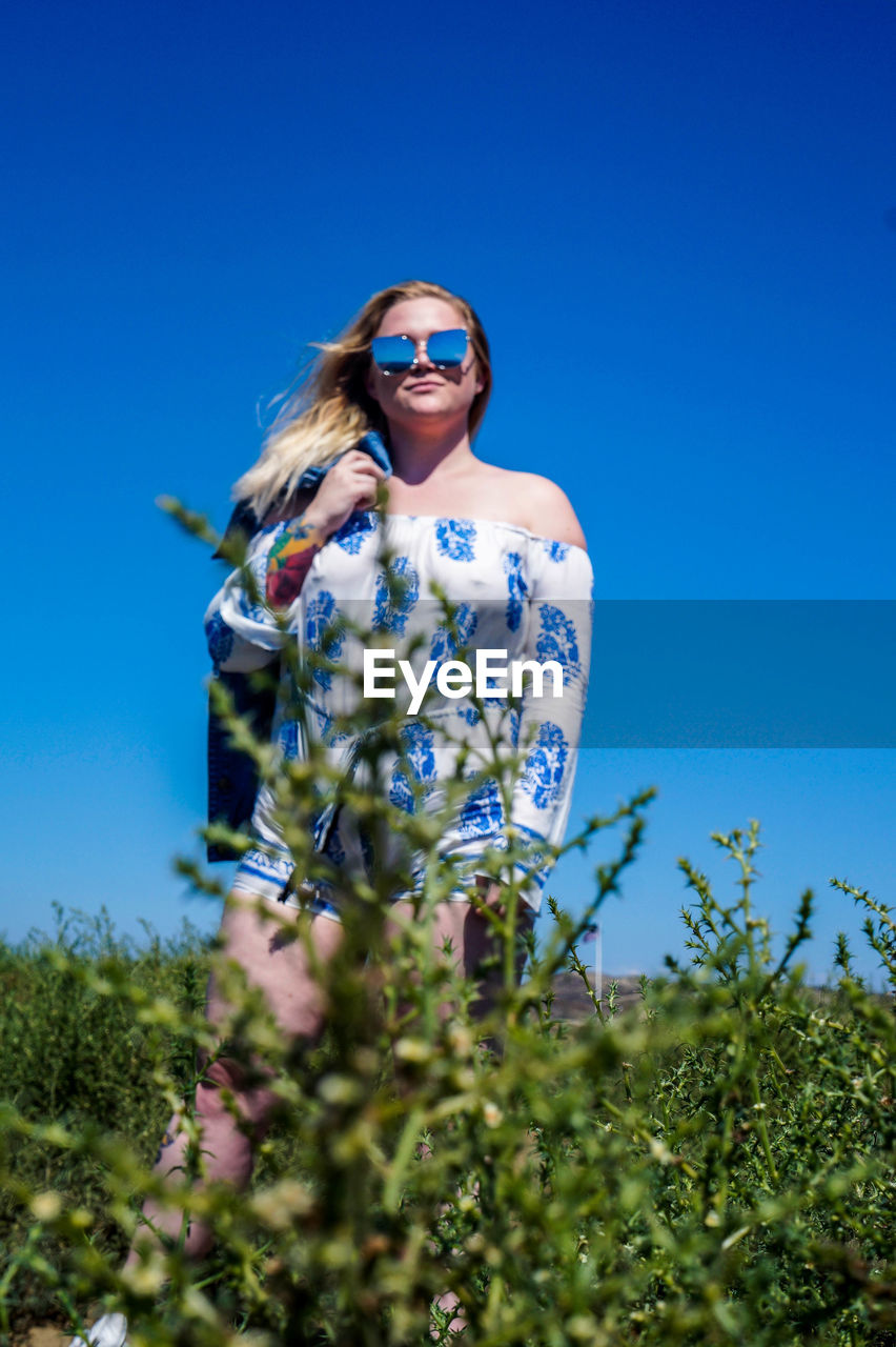 LOW ANGLE VIEW OF YOUNG WOMAN STANDING AGAINST CLEAR BLUE SKY