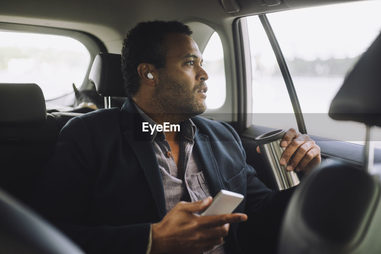 Male entrepreneur with mobile phone and insulated drink container sitting in car