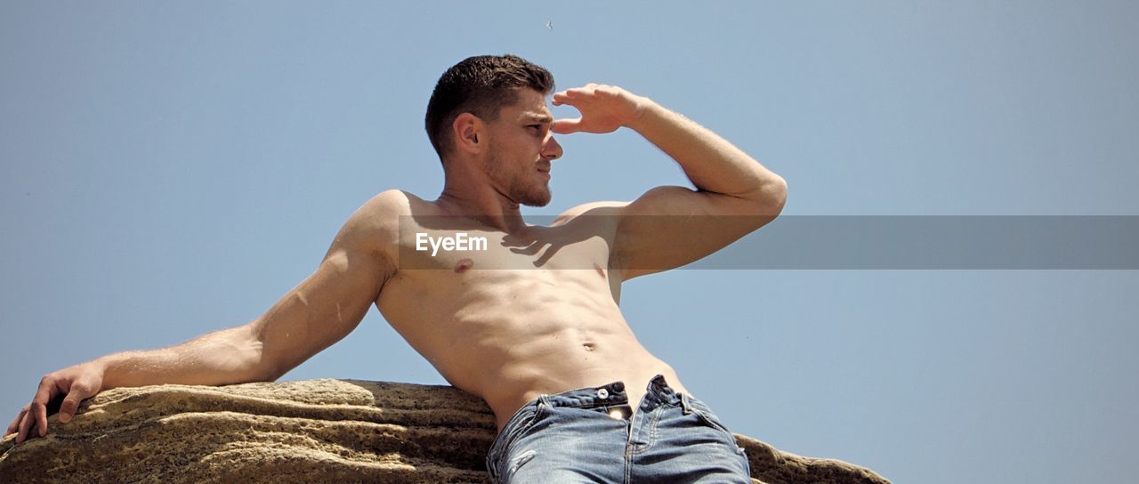 Low angle view of shirtless man shielding eyes while standing by rock against sky