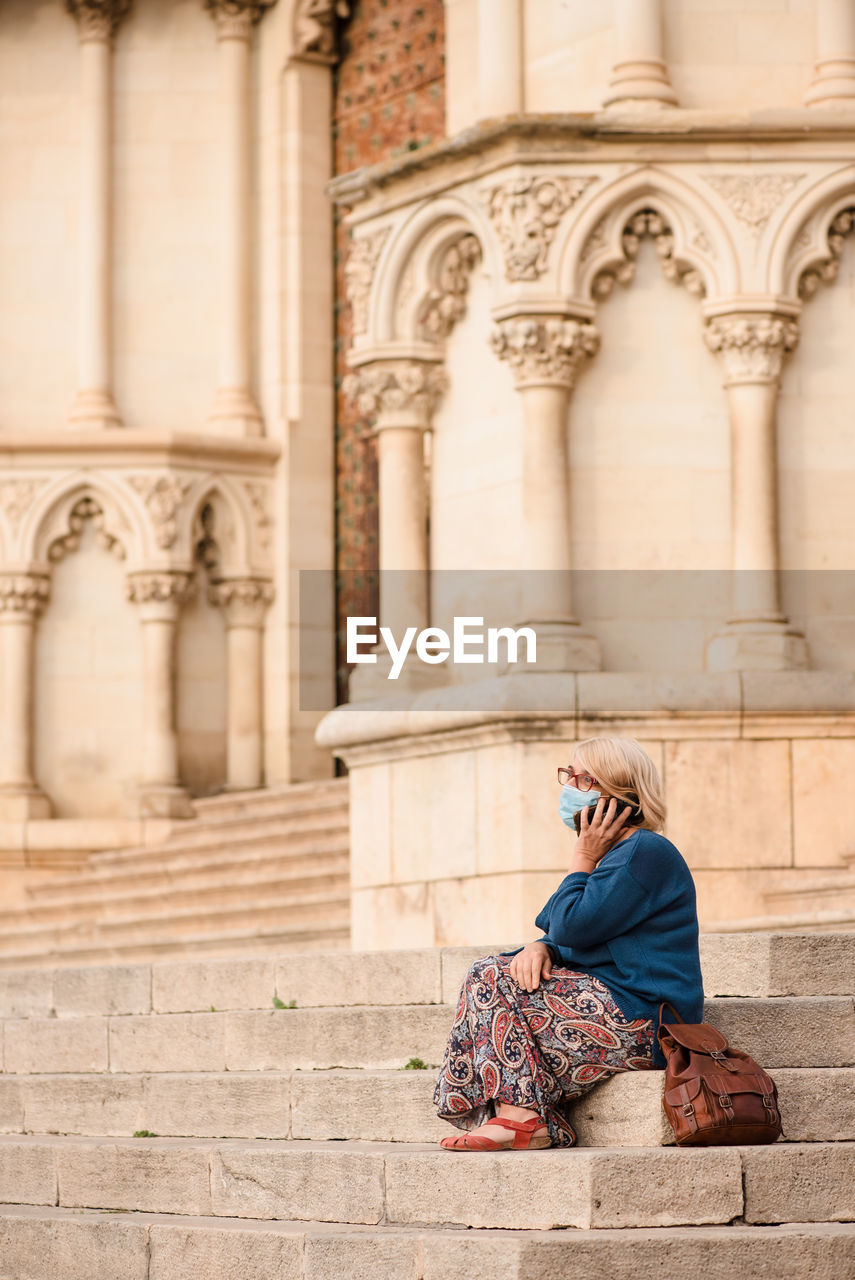 Side view of female tourist in protective mask sitting on stairs near column of medieval stone building and talking on smartphone while resting during sightseeing in cuenca town in spain
