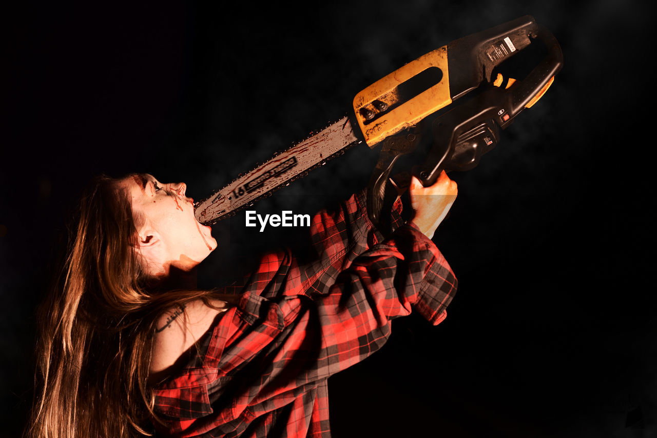 Young woman putting chainsaw in mouth at night