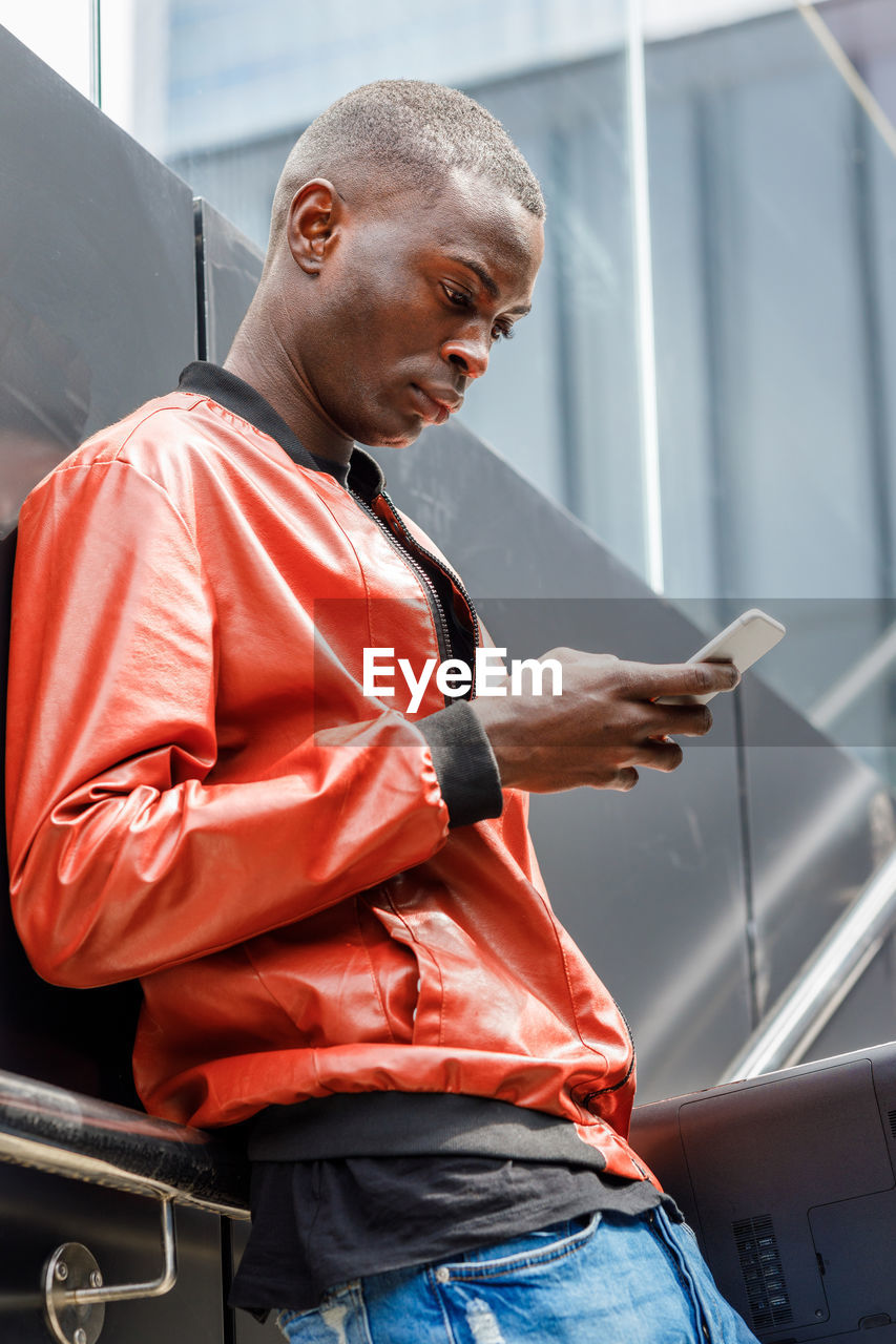 Side view of concentrated young african american guy in trendy red leather jacket messaging on mobile phone while leaning on wall on city street