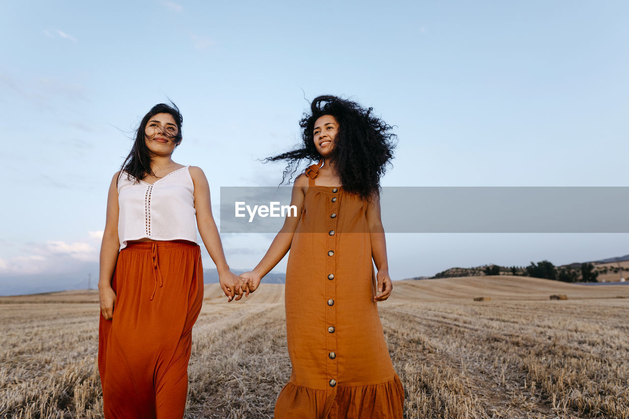 Smiling young female friends holding hands while standing in farm