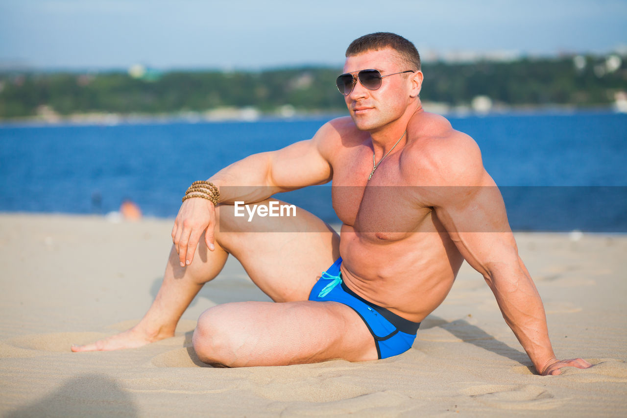 Shirtless bodybuilder while standing on beach against bay of water