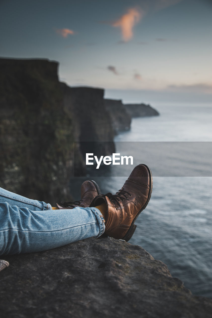 Low section of person wearing shoes on cliff by sea during sunset