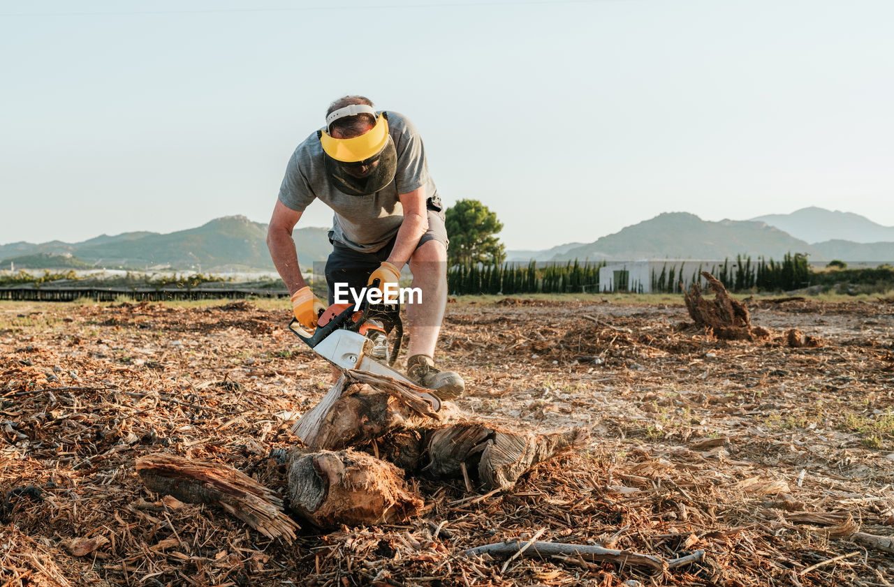 Mature unrecognizable man standing in field and starting modern chainsaw while getting ready for cutting tree trunk in rural area in highlands