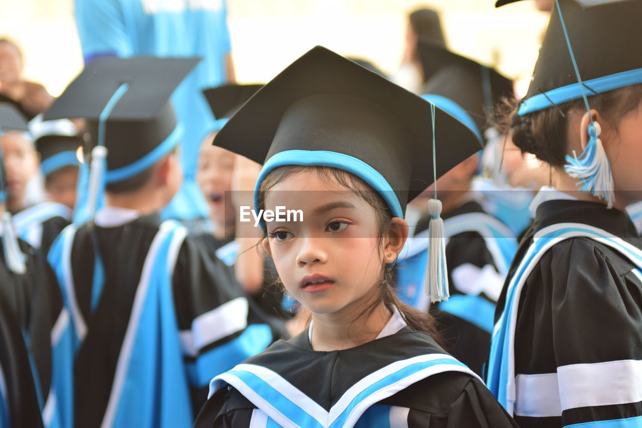 Close-up of girl wearing graduation gown and mortarboard looking away by friends